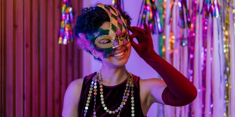 A smiling woman wearing a colorful masquerade mask and festive beads, enjoying a New Year's Eve party or carnival-themed party.