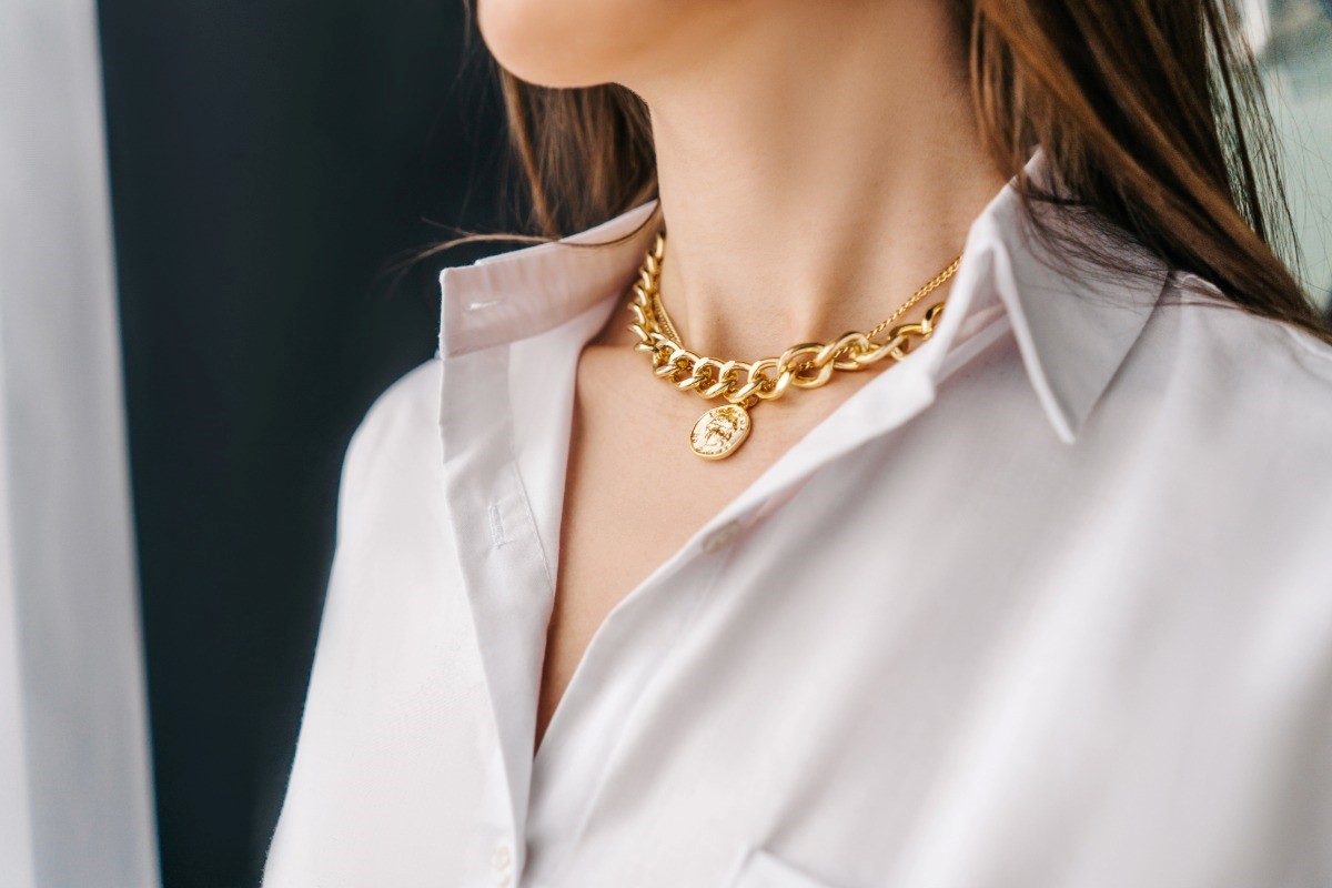 A woman wearing a stylish, layered gold-plated necklace featuring a chunky curb chain and a coin pendant over a classic white shirt.