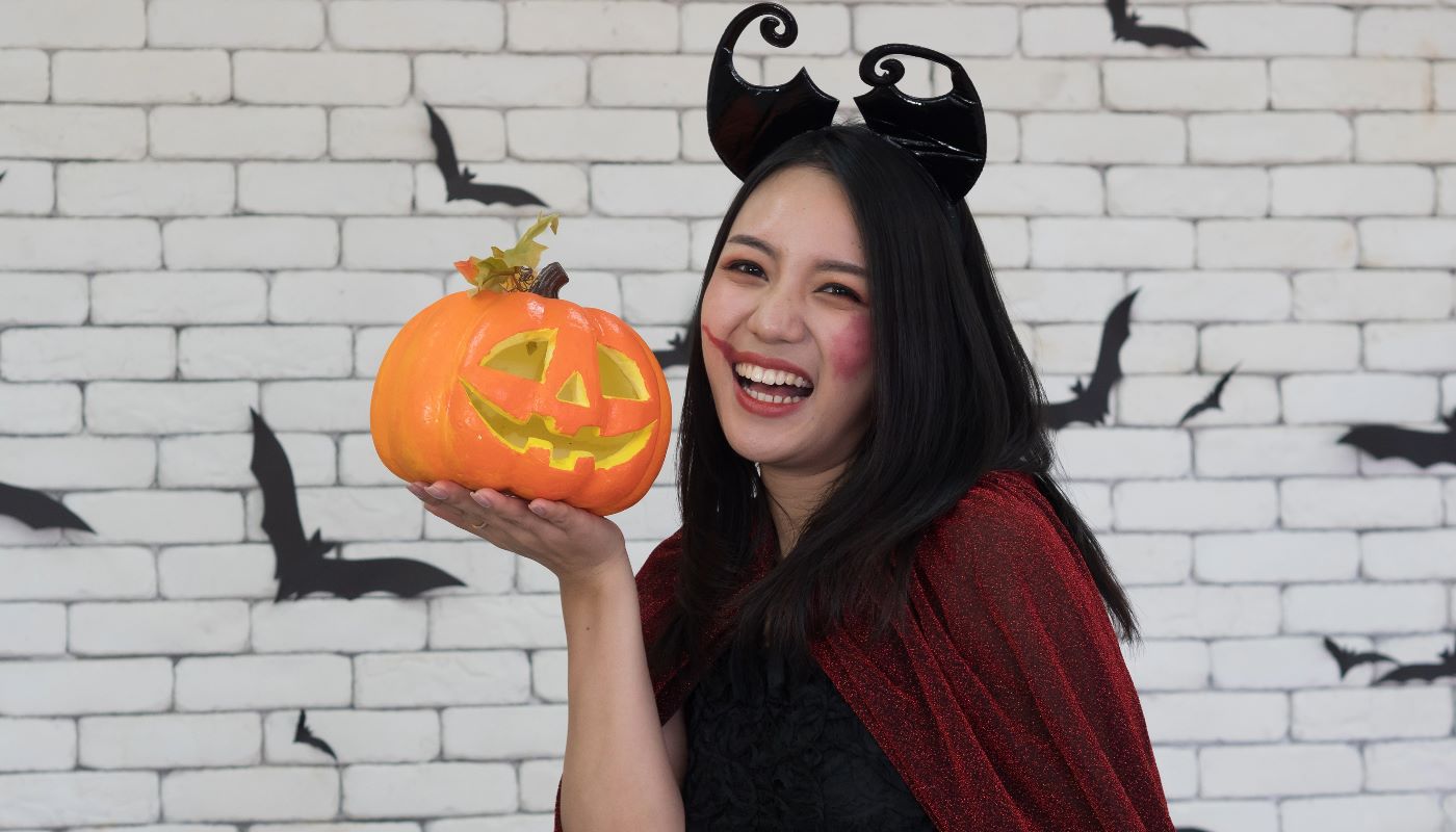 A smiling woman wearing devil horn headband, holding a carved jack-o'-lantern for Halloween.