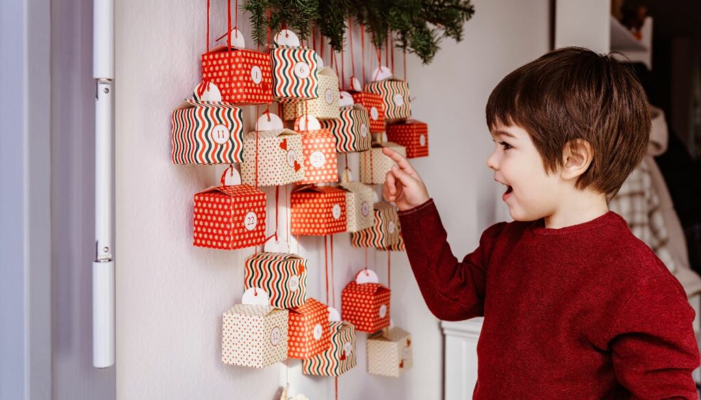 A young boy looking excitedly at a hanging DIY Christmas advent calendar made of small, decorated boxes.