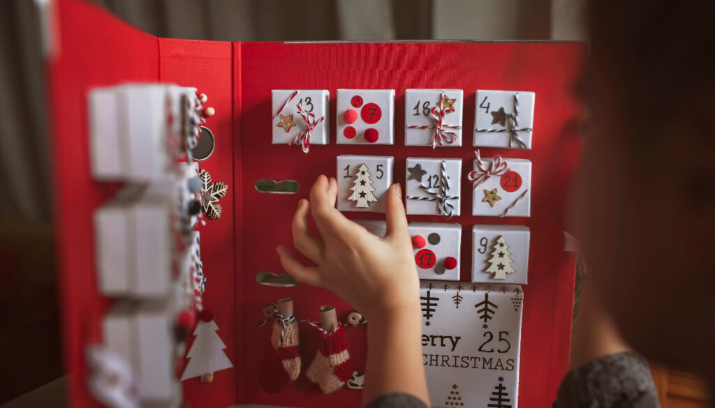 A child's hand reaching up to open a small door on a red, non-candy advent calendars for kids.