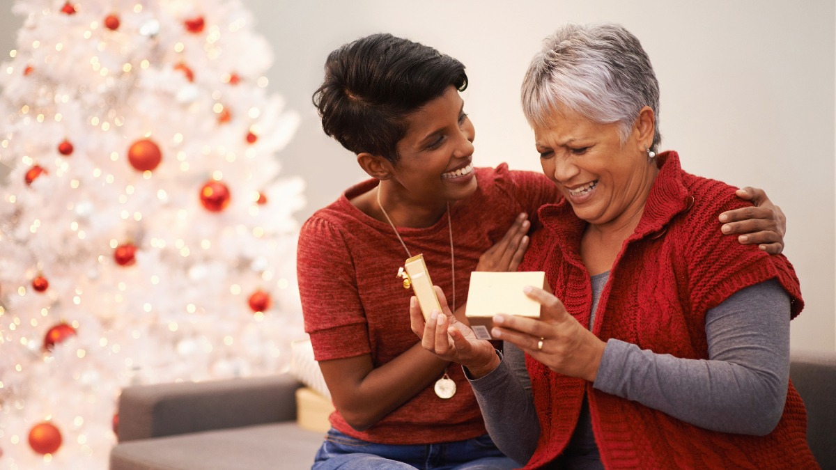 A smiling woman giving a small, wrapped Christmas gift to her mother, who is laughing joyfully.