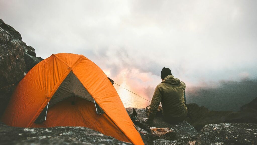 A man camping in the mountains, sitting by a campfire next to an orange tent at dusk.