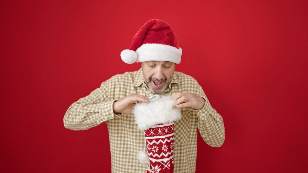 A happy father in a Santa hat looking into a Christmas stocking, as if pulling out a gift, against a red background.