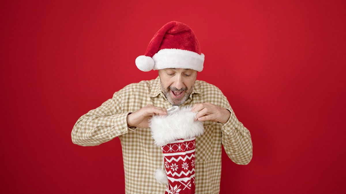 A happy father in a Santa hat looking into a Christmas stocking, as if pulling out a gift, against a red background.