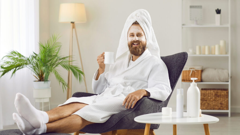 A man in a white bathrobe and a towel on his head, relaxing in a chair and enjoying a cup of coffee, representing self-care.