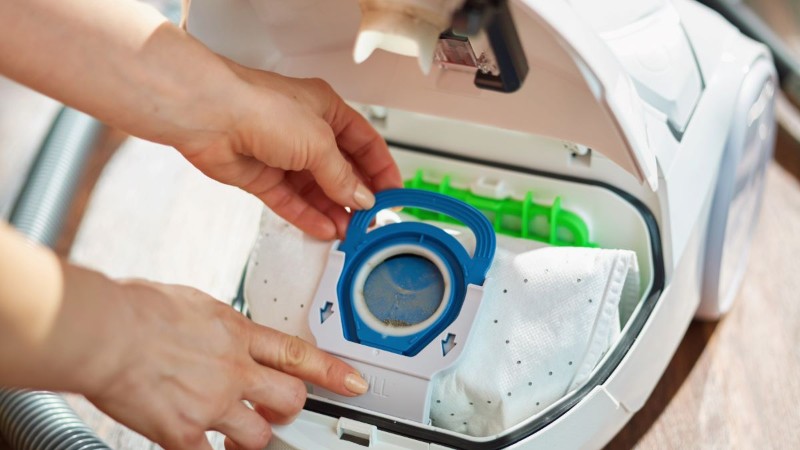 A person's hands replacing a full, disposable dust bag inside a modern canister vacuum cleaner to maintain cleaning efficiency.