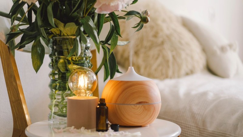 A cozy home fragrance station with a wooden essential oil diffuser, a stylish lamp with an Edison bulb, and fresh flowers in the background.