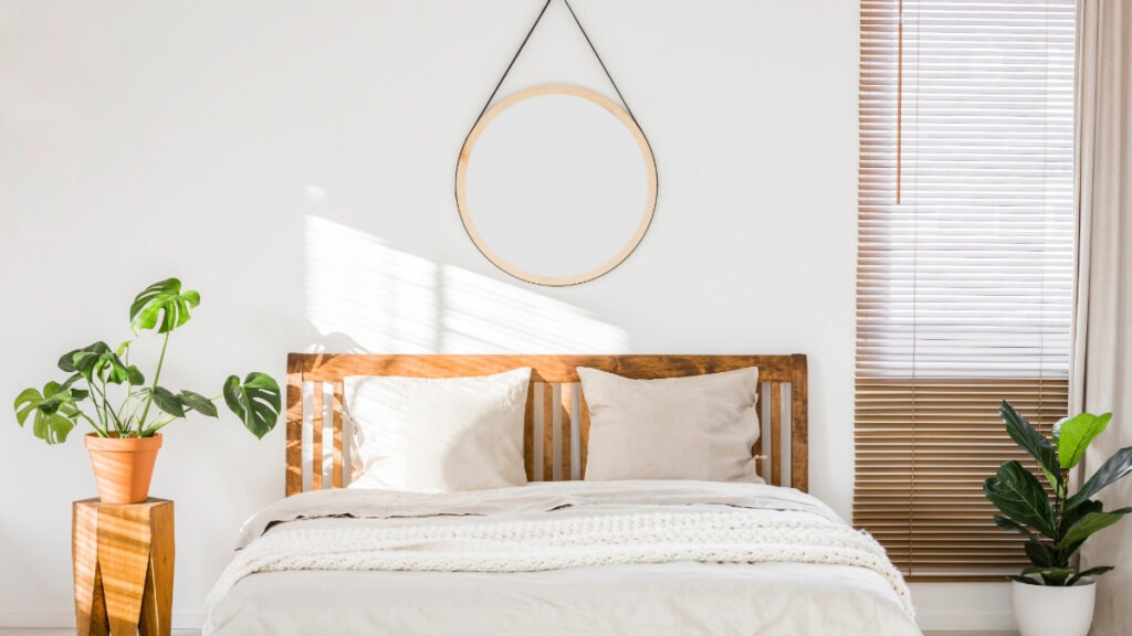 A bright and airy bedroom with a wooden headboard, white linens, scandinavian minimalism design, and a simple round mirror hanging on the wall above.
