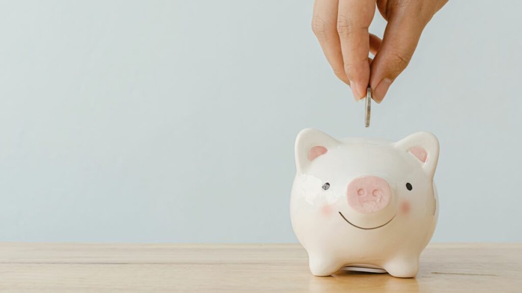 Benefits of minimalist lifestyle - saved money. A hand dropping a coin into a cute, white piggy bank, symbolizing saving money.