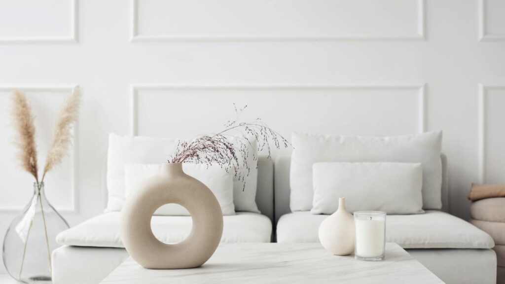An all-white, minimalist living room with a comfortable sofa and a modern, donut-shaped ceramic vase.