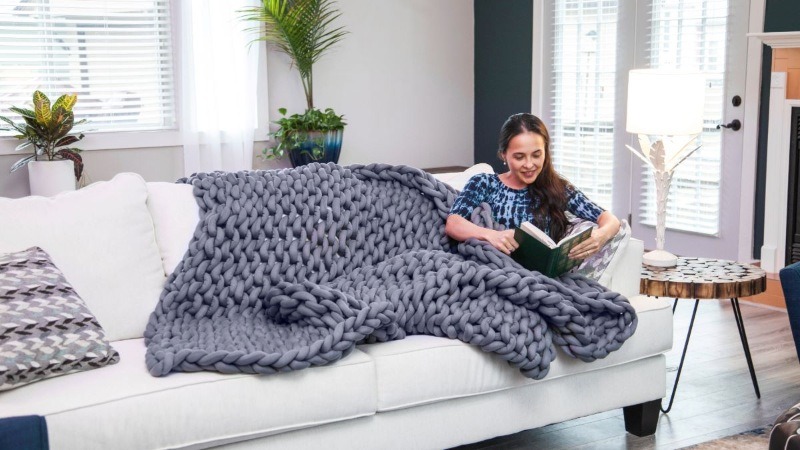 A woman reading a book while cozied up on a white sofa under a thick, grey chunky-knit blanket for ultimate comfort.