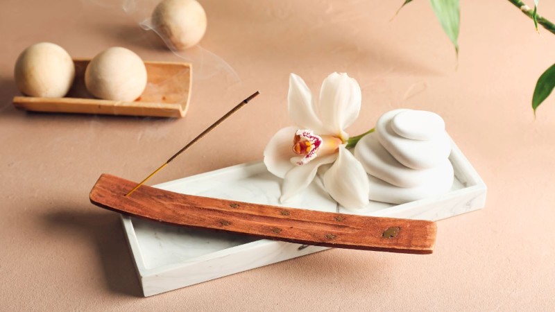 A stick of incense burning in a wooden holder, releasing fragrant smoke to make a room smell calm and amazing, next to a white orchid flower.