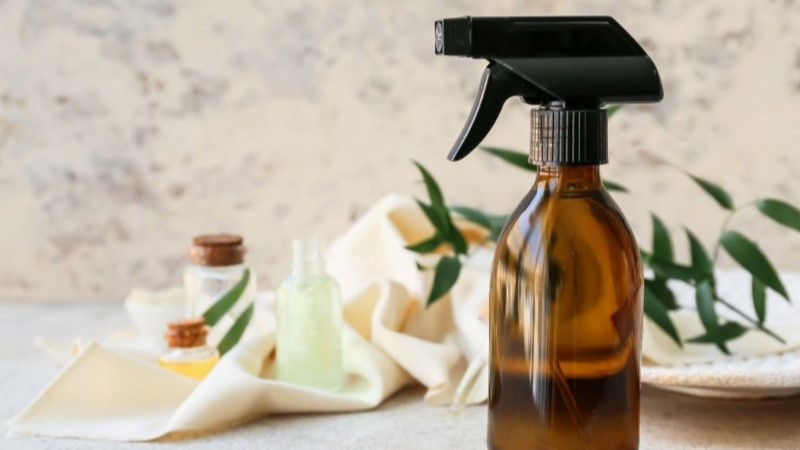 An amber glass spray bottle with essential oils and fresh green leaves, representing a natural, DIY room spray to make your home smell amazing.