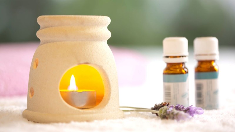 A relaxing aromatherapy setup featuring a ceramic oil burner with a lit candle, a bottle of essential oil, and a sprig of dried lavender.