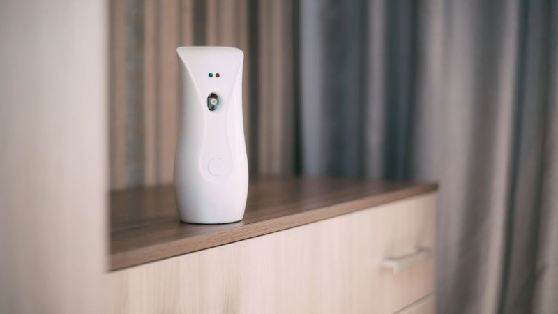 A white automatic air freshener spray dispenser sitting on a shelf, an easy way to consistently make a room smell amazing.