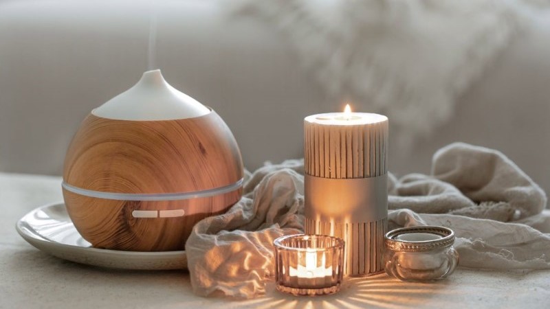 A cozy, ambient scene with a wood-grain essential oil diffuser emitting mist alongside glowing candles in a dimly lit room.