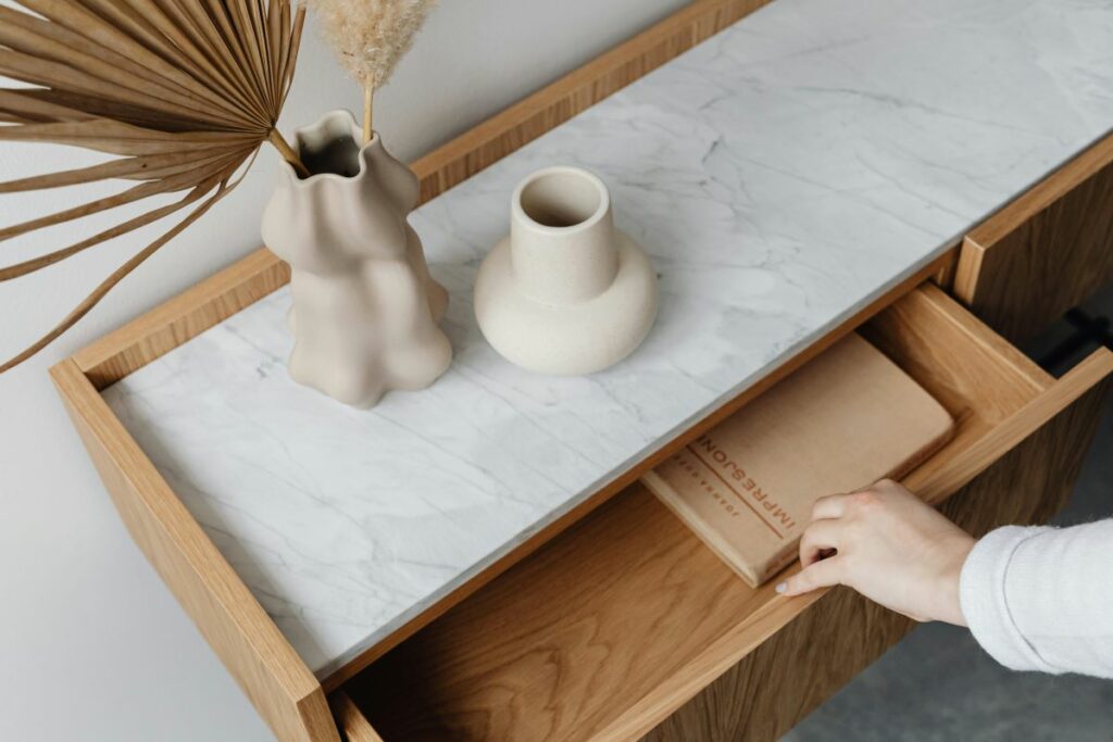 A close-up of a stylish wooden drawer with a marble top, being opened to reveal its contents, with ceramic vases on top.