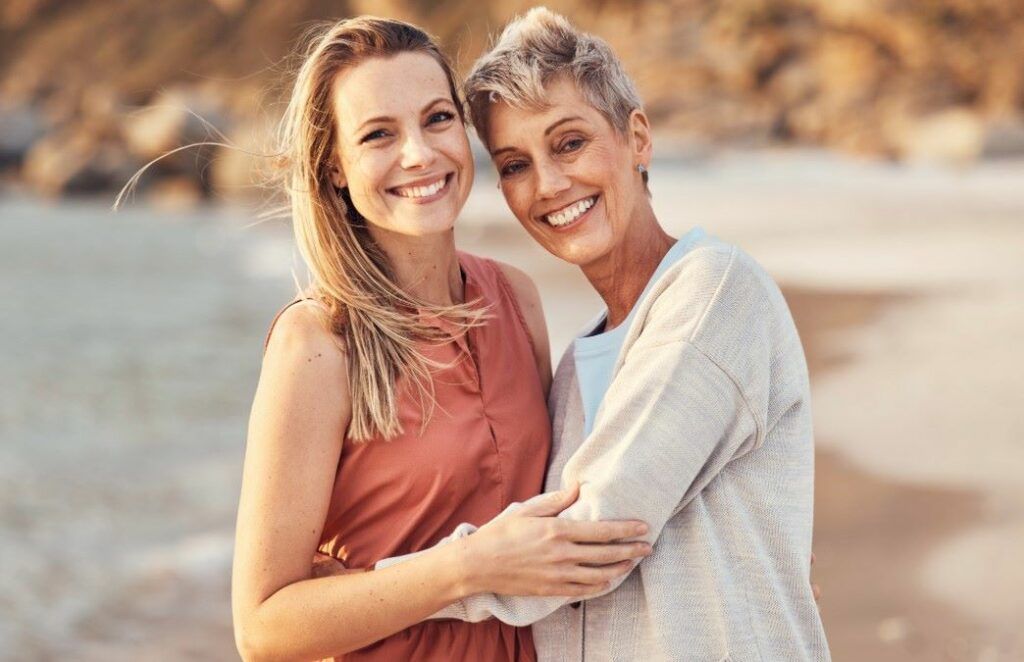 A smiling adult daughter hugging her mother warmly in an outdoor setting at sunset.