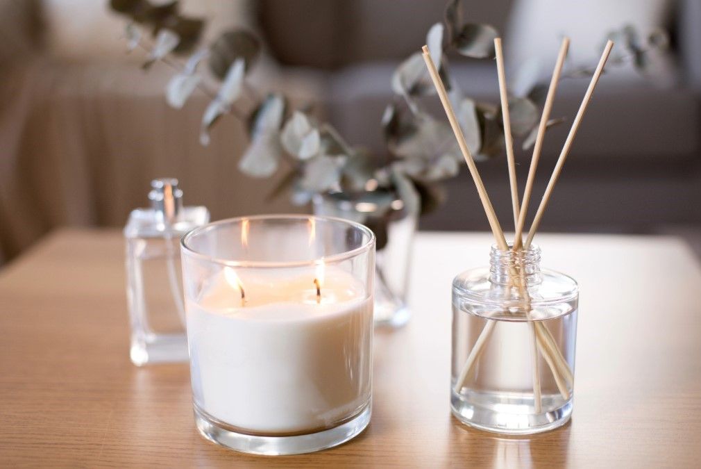 A cozy home fragrance scene with a lit scented candle, a reed diffuser, and a bottle of perfume next to eucalyptus leaves.