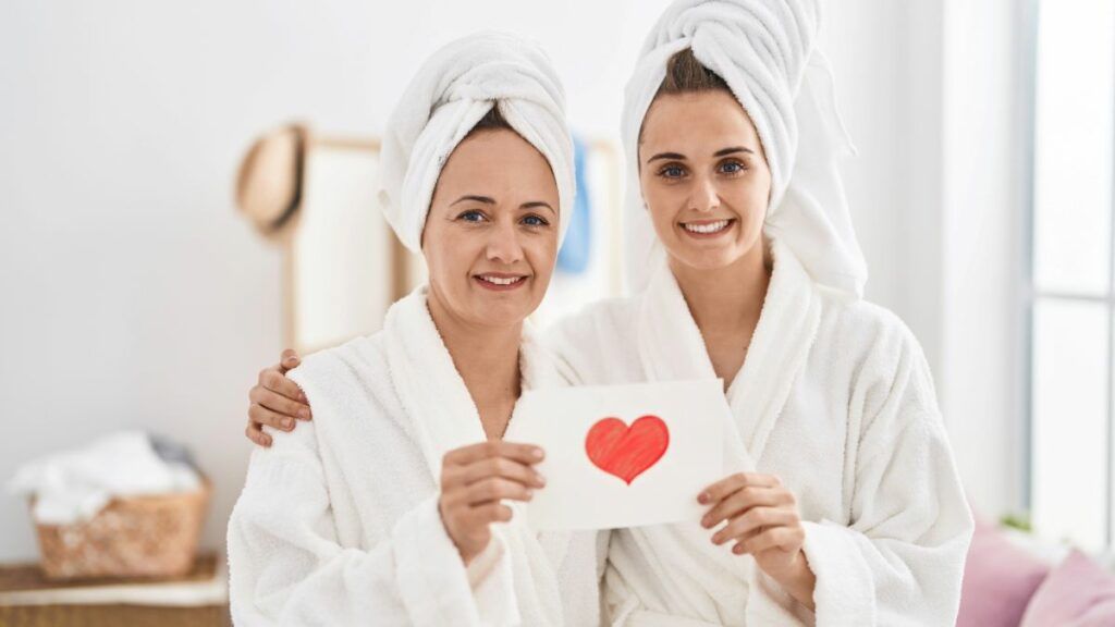 A mother and daughter wearing white bathrobes and towels on their heads, smiling and holding a card with a heart on it.