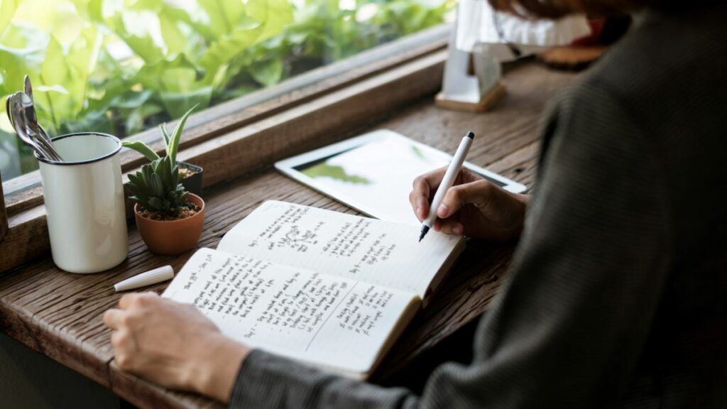 A person writing in a journal with an ink pen at a rustic wooden desk by a window.