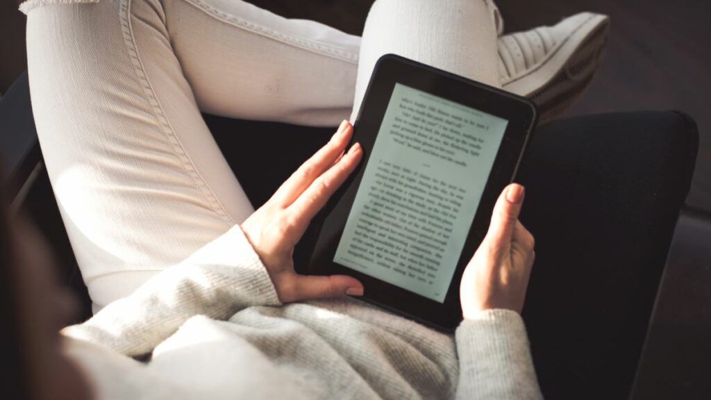 A person relaxing in a cozy chair while reading a book on an e-reader device.