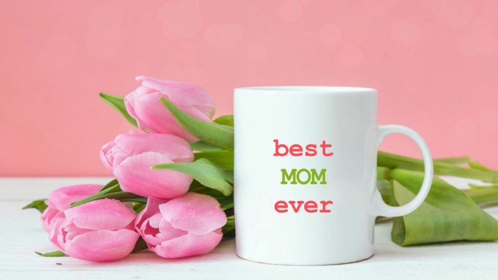 A white mug that says 'best MOM ever' in colorful letters, sitting next to a bouquet of fresh pink tulips.