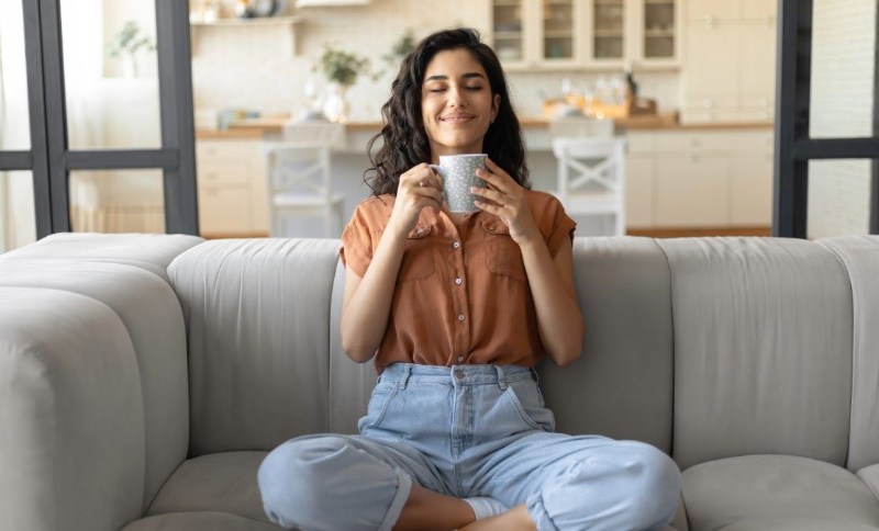 A woman relaxing on a sofa with her eyes closed, holding a warm mug in her hands and enjoying a peaceful moment at home.