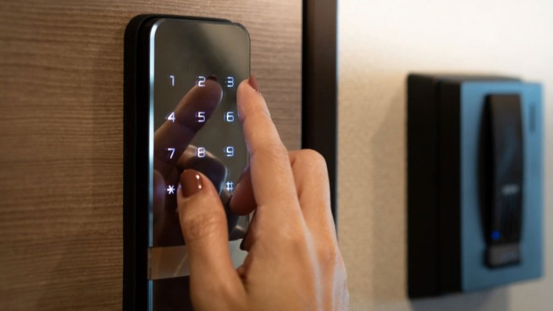 A woman's hand entering a security code on a sleek, black digital smart lock keypad, smart digital home gadgets gifts.