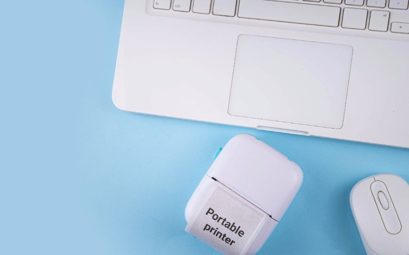 A compact, white portable printer sitting next to a laptop on a bright blue background, showcasing a convenient tech gadget for on-the-go printing.