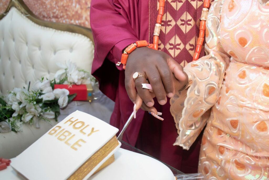 A Nigerian couple's hands, adorned with rings and coral beads, resting near a Holy Bible during their traditional wedding ceremony.
