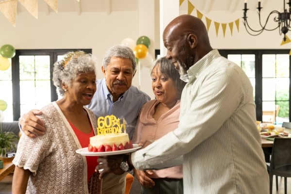 A senior woman with grey hair smiles joyfully as her loved ones present her with a cake to celebrate her 60th birthday, representing a perfect gift moment for a woman in her 60s.