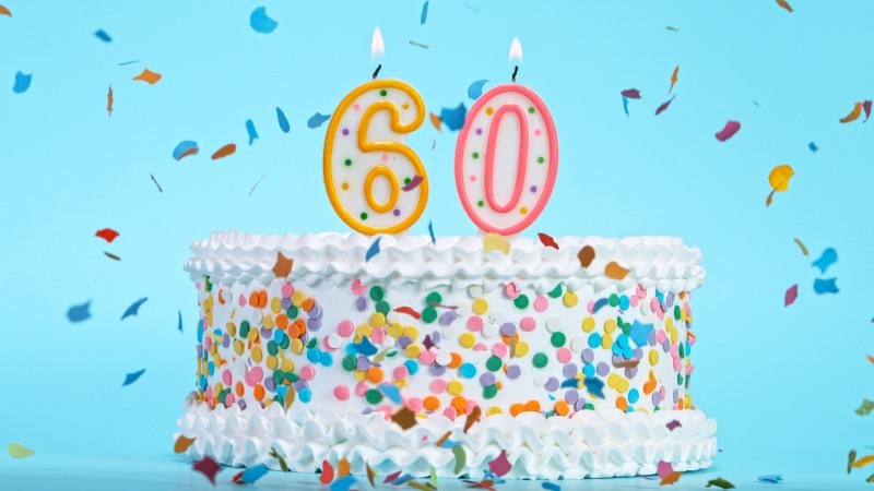 A festive 60th birthday cake for women with colorful sprinkles and lit candles shaped like the number 60, with confetti falling against a blue background.