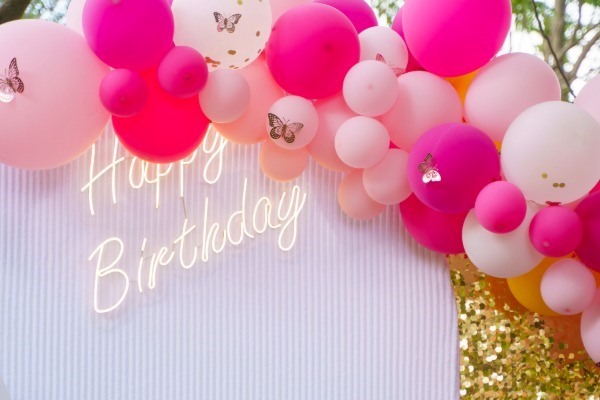 A stylish birthday party backdrop featuring a large balloon garland with shades of hot pink and light pink balloons. A white neon sign that says 'Happy Birthday' glows against a white ribbed wall.