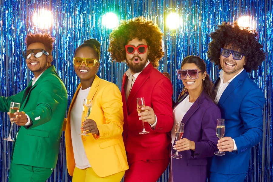 Guests dressed in colorful suits and afro wigs for a creative 70s disco party theme, holding drinks and posing in front of a glittery backdrop.