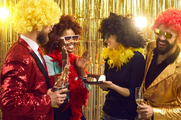 Friends enjoying a costume party, wearing colorful wigs and feather boas while laughing and sharing cake in front of a festive gold backdrop.