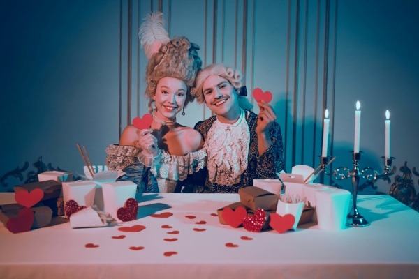 Two guests dressed in elaborate 18th-century historical costumes and powdered wigs, enjoying a creative themed dinner party.