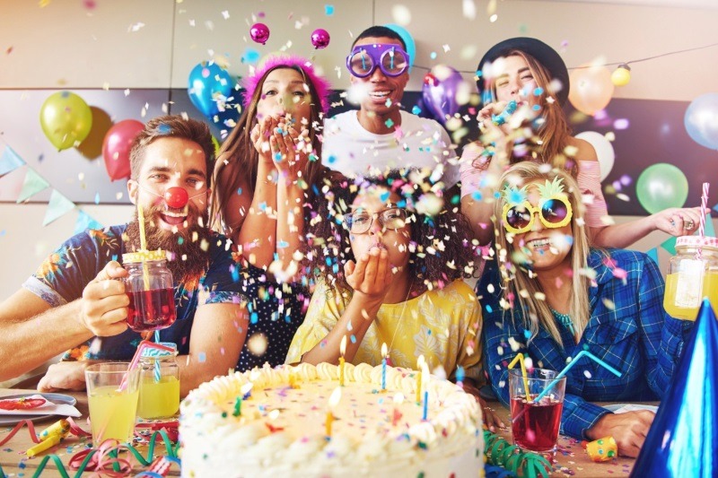 A lively birthday party celebration where a diverse group of friends in silly glasses blow confetti over a birthday cake with lit candles.