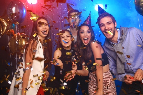 A group of friends enjoys a high-energy party, laughing under a shower of golden confetti while wearing festive party hats.