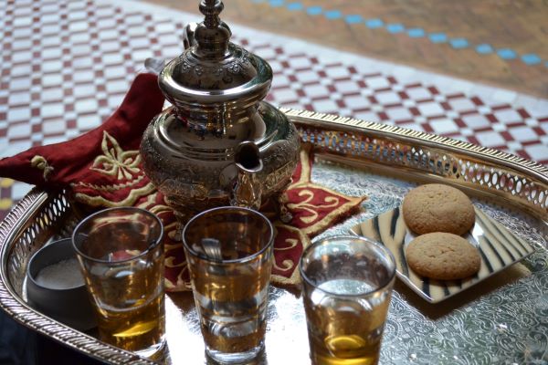 An engraved metal Moroccan tea set, including an ornate teapot and glass, arranged on a decorative tray table with a carved wooden base.