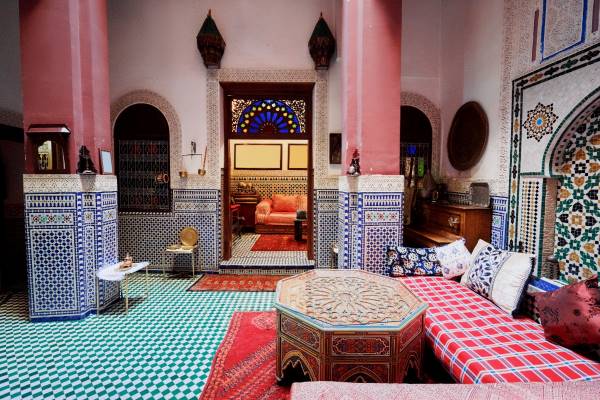 A bright and airy Moroccan-style living room featuring a large, ornate archway, a low wooden coffee table, and a colorful patterned rug. A traditional metal lantern hangs from the ceiling.