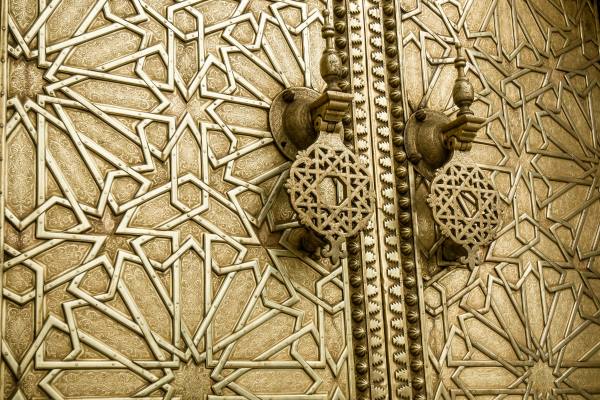 A pair of ornate, antique Moroccan wooden doors featuring intricate geometric patterns in faded colors, set within a beautifully tiled archway.