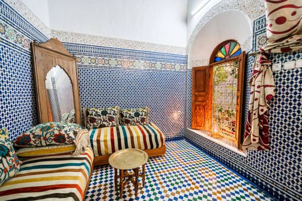 A serene Moroccan living room featuring two colorful cushioned sofas, a large mirror with a wooden frame, walls with Morrocan tiles, and small table in the middle.